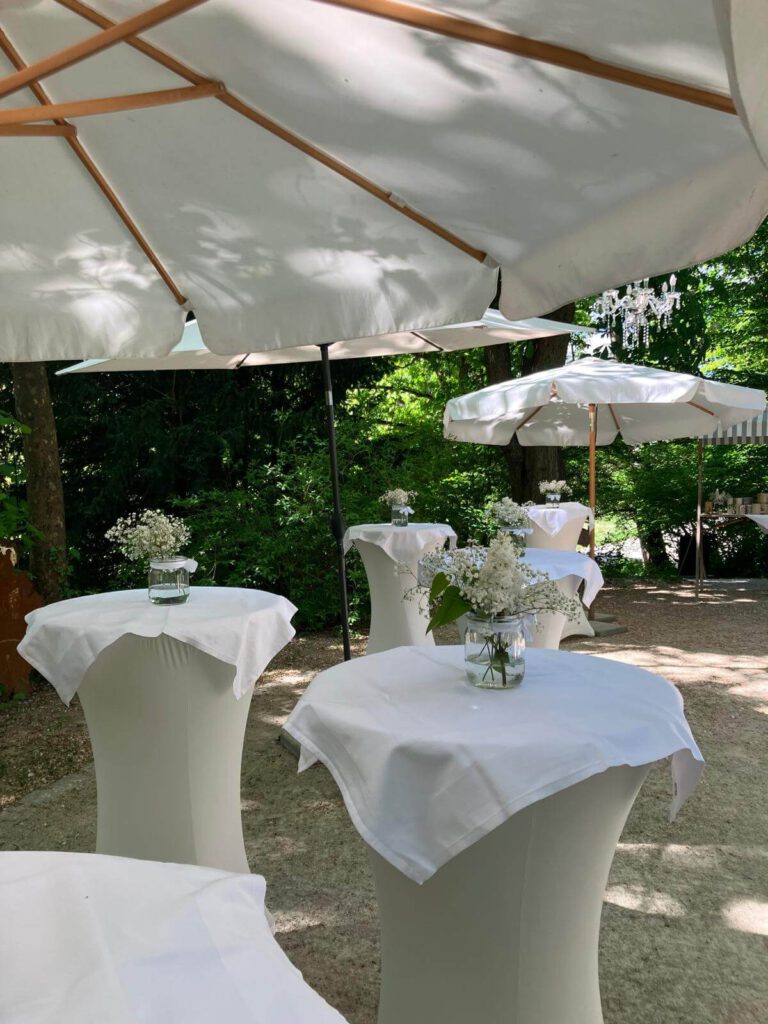 Stehtische mit weißen Tischtüchern und Blumen unter Sonnenschirmen im Garten